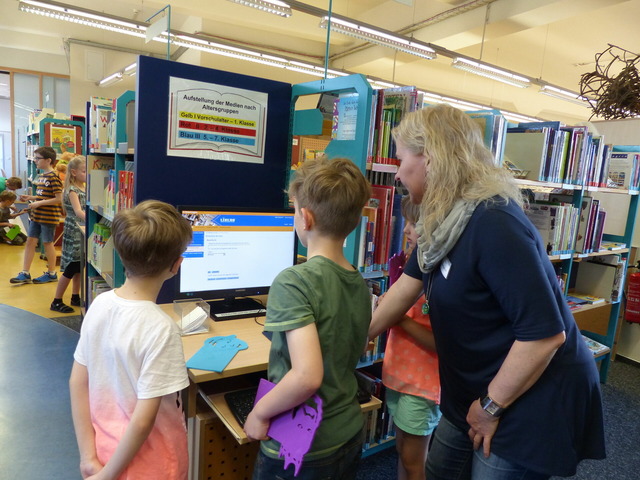 Das Bild zeigt zwei Kinder und eine Bibliothekarin am Computer.